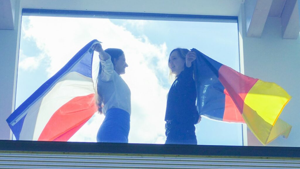 Zwei Frauen stehen sich gegenüber und schauen sich an, eine von ihnen hält eine Deutschlandflagge hoch und die andere eine Frankreichflagge